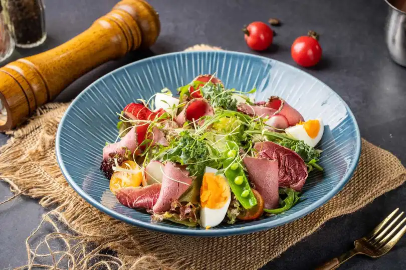 旁边放着一份带有肉和蔬菜的沙拉盘 旁边还放着一碗番茄汤。