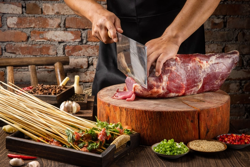 一个人正在一块木板上切肉 木板上还放着香料和蔬菜 桌子上有一把大刀和新鲜食材。