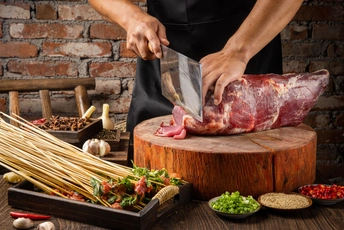 一个人正在一块木板上切肉 木板上还放着香料和蔬菜 桌子上有一把大刀和新鲜食材。