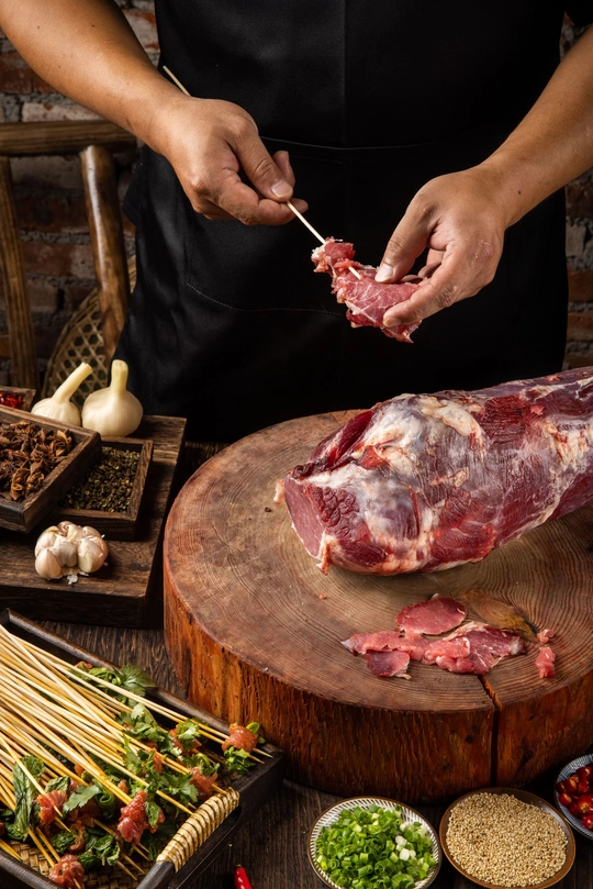 一个人在木板上切着羊肉 木桌上还放着其他配料和蔬菜。