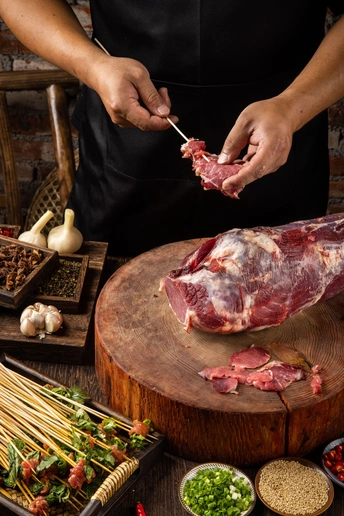 一个人在木板上切着羊肉 木桌上还放着其他配料和蔬菜。