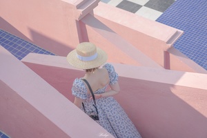 一位穿着蓝色连衣裙和草编帽子的女人坐在粉红色瓷砖墙上