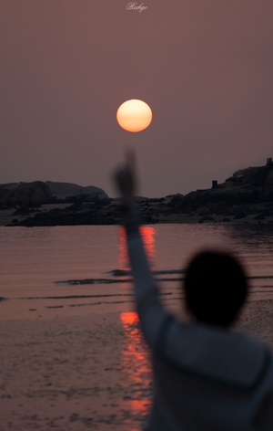 一个人举起手臂在水上拍摄夕阳西下 天空中满月升起。