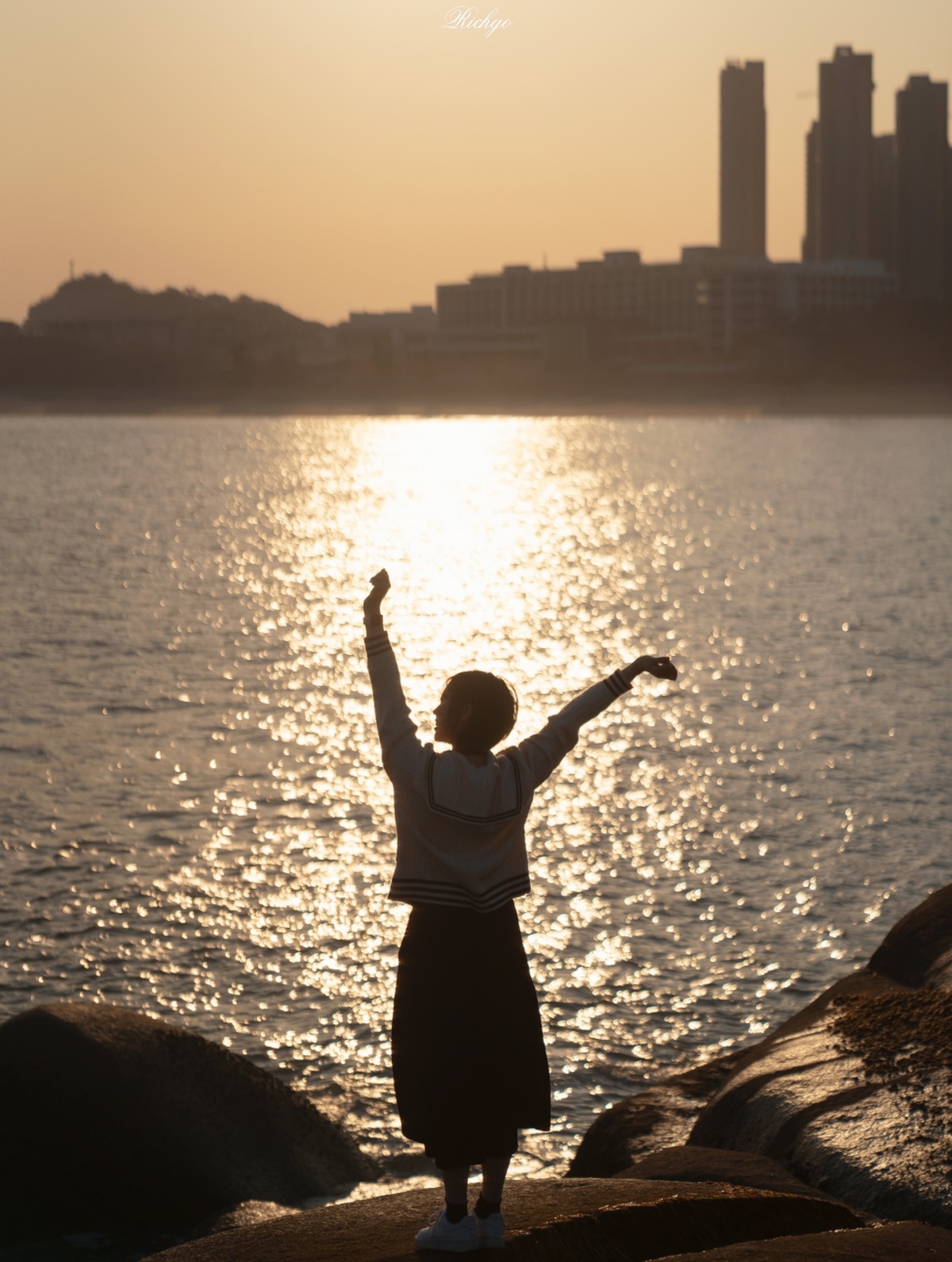 一个年轻女子站在夕阳下 前方有一片水域 她伸展双臂 手中拿着一只风筝。