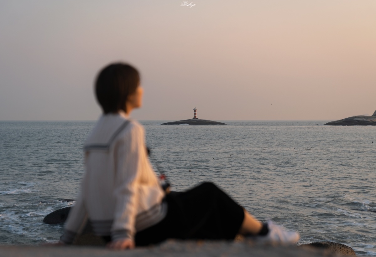 一个年轻男子和女子坐在岩石上 眺望着落日 远处有一座岛屿和一座灯塔。