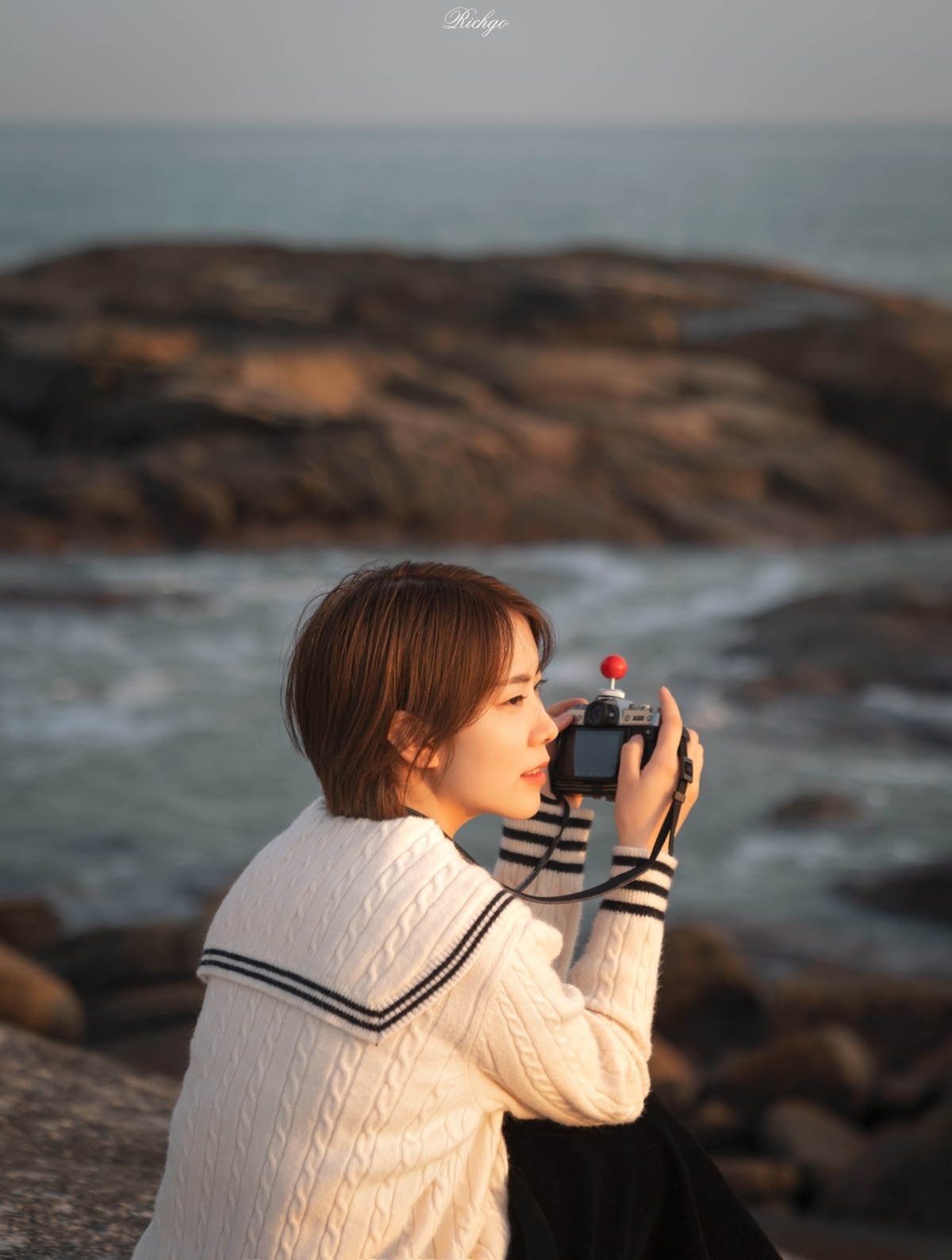 一位年轻女子手持相机 正在拍摄前景为岩石的海或海景。