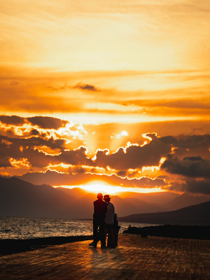 夕阳下 一对情侣在码头拥抱 夕阳在水面上落下 人们在沙滩上站着。