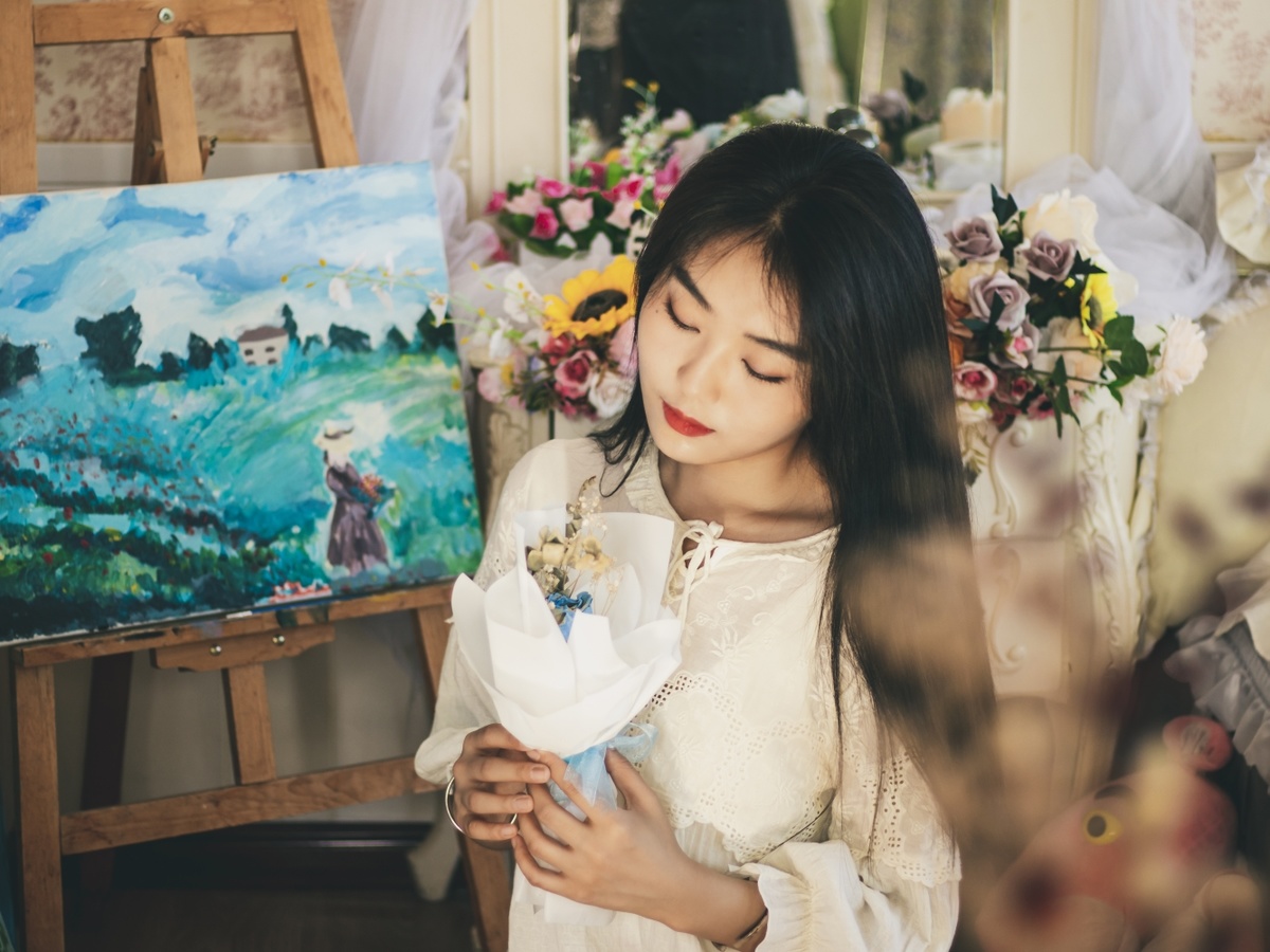一个年轻的女人拿着一朵白花站在一个工作室里的艺术家的照片前 工作室里有绘画和画布