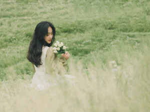 一个穿着白裙子的年轻女子坐在一片高草地上 闻着一朵花。