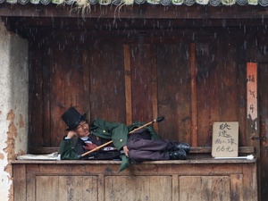 一些人正在一所建筑物的外面 在雨中睡觉 窗户是木制的 一个人躺在阳台上 手里拿着一把伞。