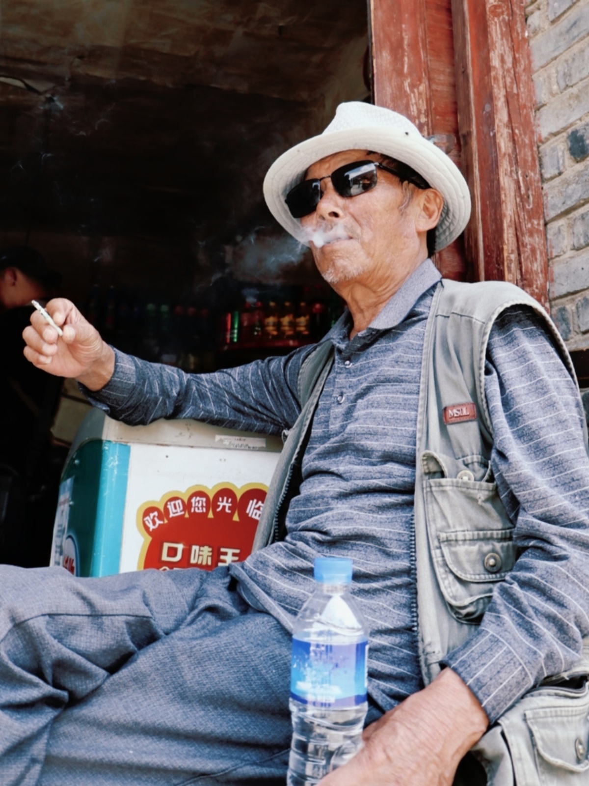 一个戴帽子、太阳镜、抽烟、坐在建筑物前、手拿一瓶水的老人。