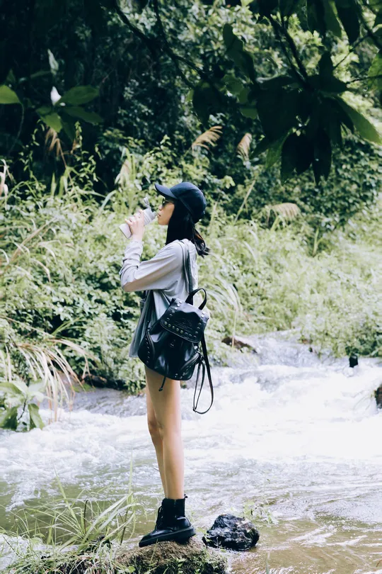 一位年轻女子手持相机 正在为一只站在岩石上的鸟儿拍照 该岩石位于森林中的一条溪流中。