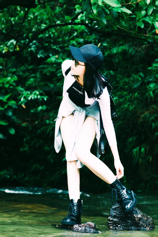 一位穿着黑色靴子和帽子、站在溪流中的岩石上、手持雨伞的年轻女子。