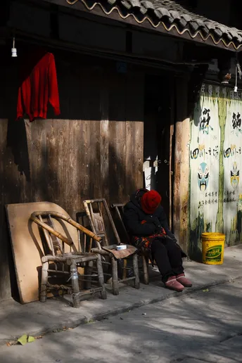 一位穿红衣服的女人坐在一条木制摇椅上 摇椅位于一幢老建筑前的人行道上。