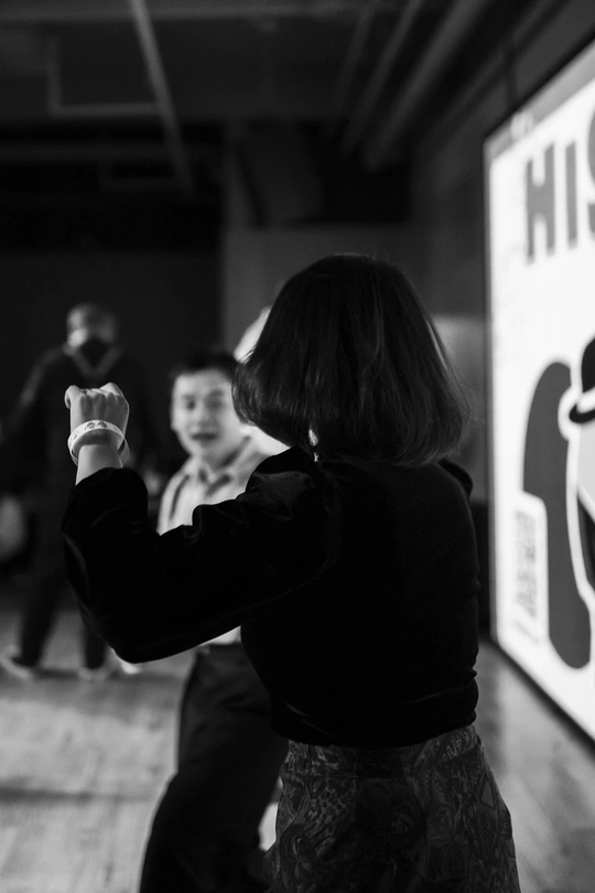 一张黑白照片 一个年轻女孩在和其他人一起玩电子游戏 周围站着其他人。