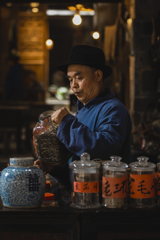 一个戴蓝帽子的老人坐在有其他茶叶罐的桌子旁 手里拿着一罐玻璃瓶装的茶。