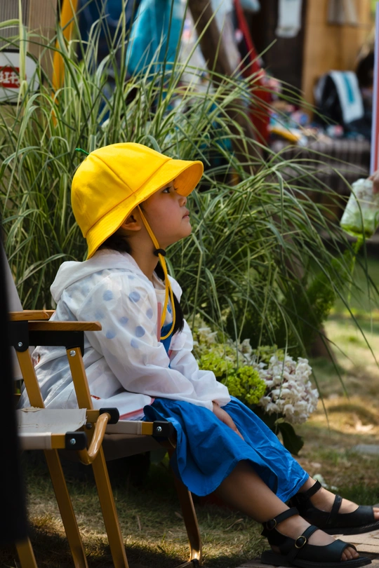 一个小女孩戴着黄帽子坐在一个小木凳上 一个小男孩戴着黄帽子坐在遮阳伞下。