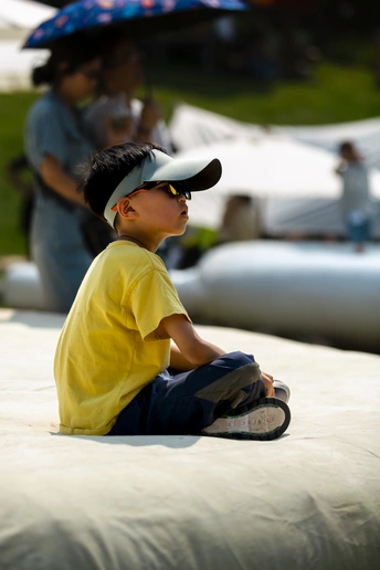 一个穿着黄色衬衫和戴着帽子的小孩子坐在地上 背景中有把雨伞。