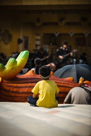 一个穿着黄色衬衫的男孩坐在一排船旁 背景中有人 还有一个穿黄色衬衫的男人。