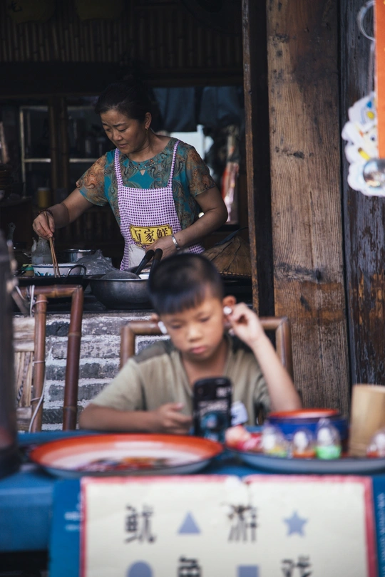 一个年轻男孩坐在桌子旁 一个妇女站在他身后 对着手机在前面的餐厅里讲话。