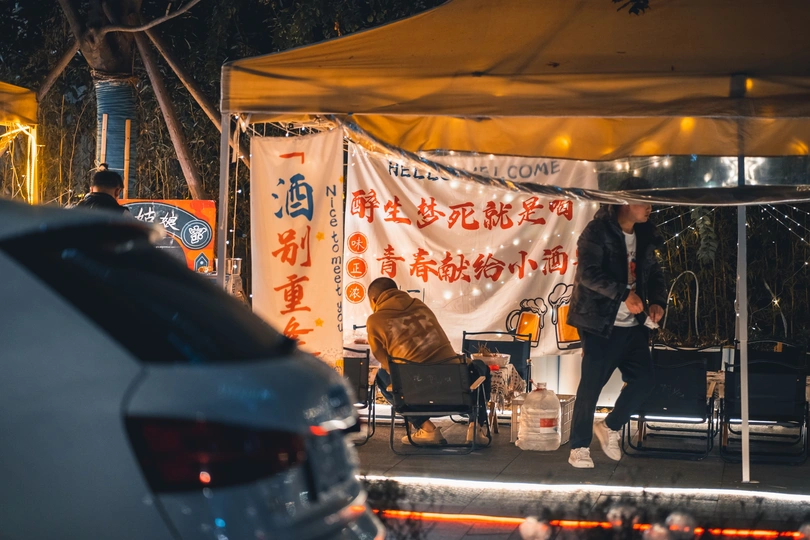 夜晚街道上有一顶帐篷 一个人站在一辆车旁边 狗在车上坐着。