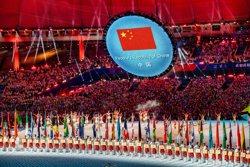 一张在大型体育场举行的体育赛事上 有众多运动员和表演者的大型群众照片 舞台上飘扬着奥林匹克旗帜。