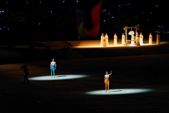 一位女性站在夜晚发光的舞台上 背景有其他舞者和表演者。