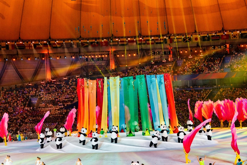 许多人聚集在一个大体育馆里 体育馆中央有五颜六色的旗帜 为一场表演做准备。