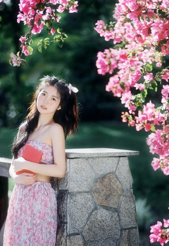 一位穿着美丽裙子的年轻女子 手里拿着一本书 站在一个有粉色花朵的公园里。