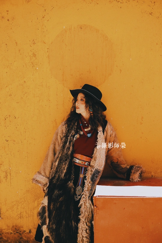 一个戴黑色帽子、有毛领的外套的女孩站在黄色墙壁前