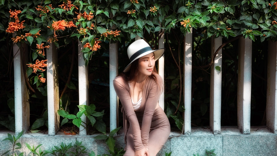 一位穿着连衣裙和白色帽子 坐在长凳上 靠在有花墙上的年轻女子