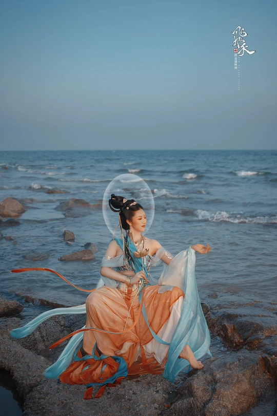 一位穿着蓝色连衣裙的美女坐在海边岩石上吹泡泡。