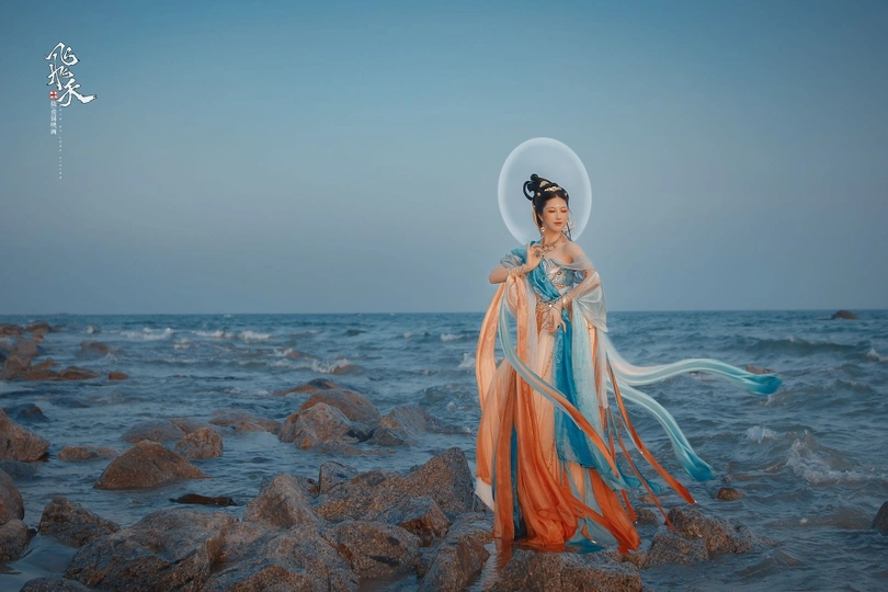 一位穿着蓝色连衣裙的美女站在岩石上 岩石位于海洋边的岩石海滩上 背景中是月亮。