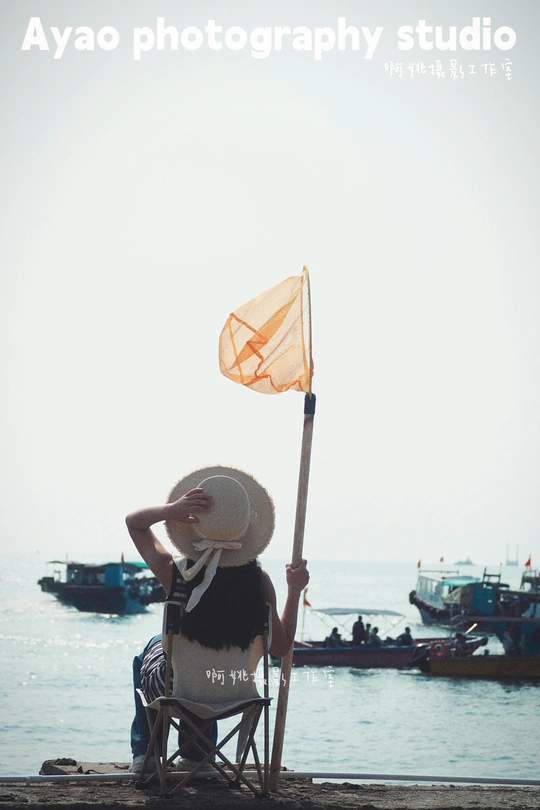 一位女性坐在椅子上拿着一把伞和一面旗 与一位戴着帽子的男性坐在海滩上 背景中有海洋中的船。