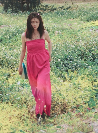 一位穿着粉红色连衣裙 手持雨伞 穿过花丛和草地的年轻女子。