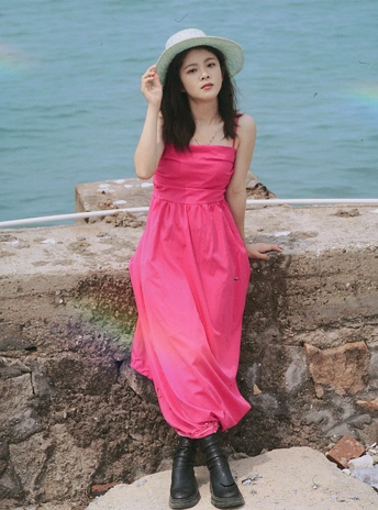 一位穿着粉色连衣裙和靴子的年轻女子站在一块石头墙上 俯瞰着一片水域和一片海。
