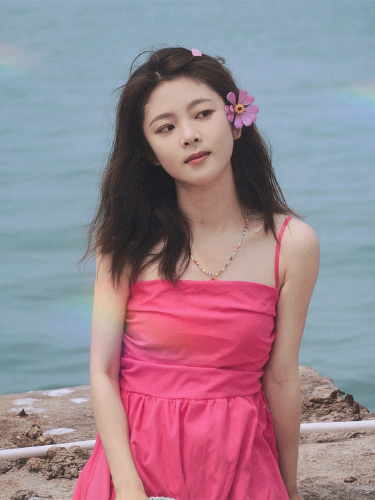 一位穿着粉红色连衣裙 头戴花的女青年坐在靠近水域的海滩上。