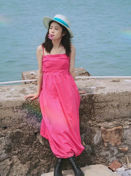 一位穿着粉色连衣裙和靴子的年轻女子在靠近水域的石头墙上摆姿势 背景是海滩。