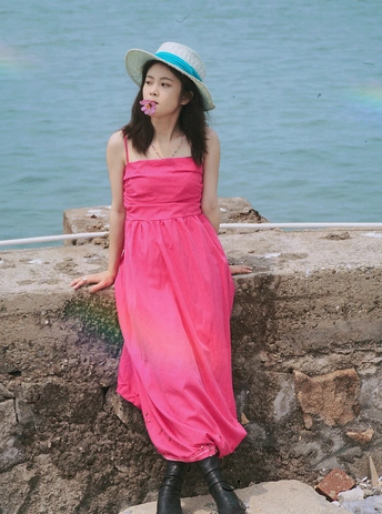 一位穿着粉色连衣裙和靴子的年轻女子在靠近水域的石头墙上摆姿势 背景是海滩。