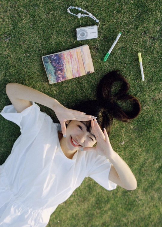 一位穿着白色连衣裙的年轻女子躺在草地上 为拍照而摆姿势 手中持有一台相机。