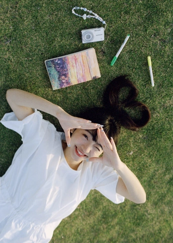 一位穿着白色连衣裙的年轻女子躺在草地上 为拍照而摆姿势 手中持有一台相机。