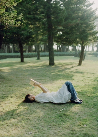一个年轻女子躺在公园的草地上阅读一本书 背景中有树木和一个男子躺在那里。