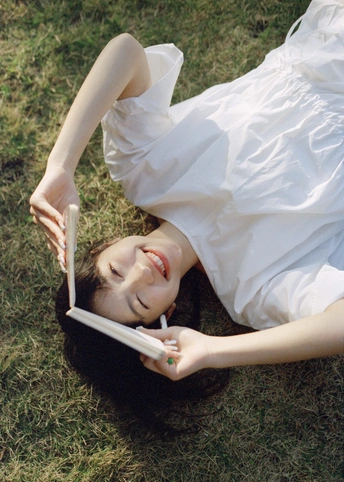 一个穿着白色连衣裙的年轻女子躺在草地上 手里拿着书和平板电脑。