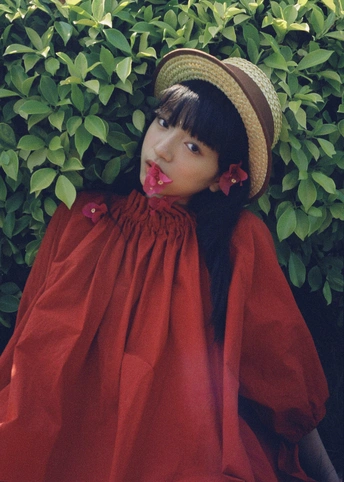 一位穿着红色连衣裙和草帽的年轻女子站在绿叶丛生的灌木前摆姿势