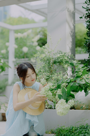 一个穿着蓝色连衣裙的年轻女子 戴着草编帽子 坐在温室前面的地上 身后是植物。