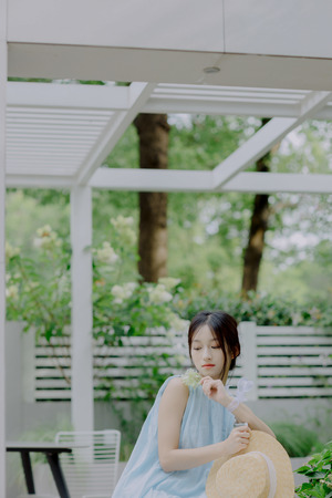 一位穿着蓝色连衣裙的年轻女子 手里拿着一朵花和一顶草帽 坐在花园里的长椅上。