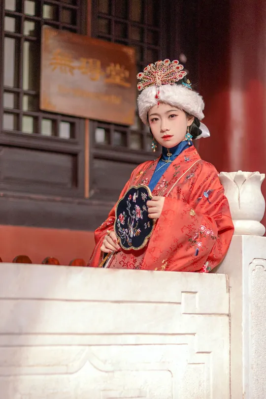 一位穿着传统红白相间服装的年轻女子站在一栋建筑前 手里抱着一个人。