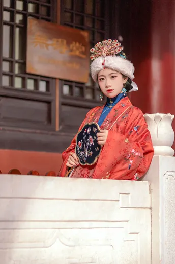 一位穿着传统红白相间服装的年轻女子站在一栋建筑前 手里抱着一个人。