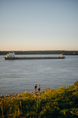 夕阳下 几人在河岸边行走 远处有一艘大型驳船。