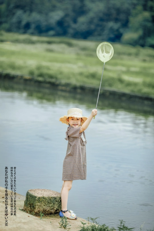 一个穿着连衣裙、戴着帽子的年轻女孩站在湖边或河边的岩石上 手里拿着一个蝴蝶形状的气球